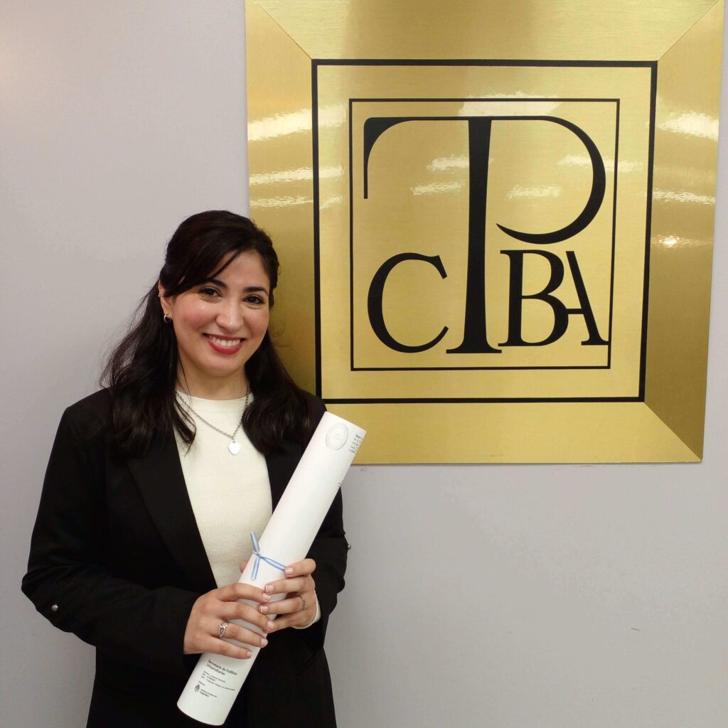 traductor-pública-matriculada-Luciana-Gomez_CTPCBA Luciana Gomez, traductora pública de inglés, sosteniendo un diploma enrollado frente a un letrero del CTPCBA.